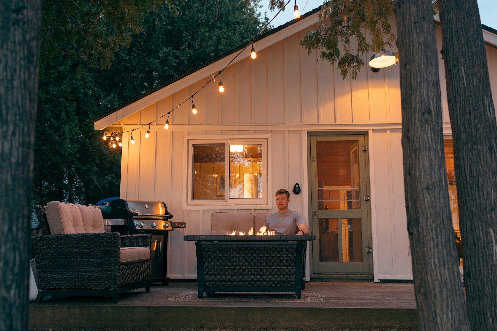 Cozy cabin porch at dusk with string lights, wicker chairs, and a person seated at a table.