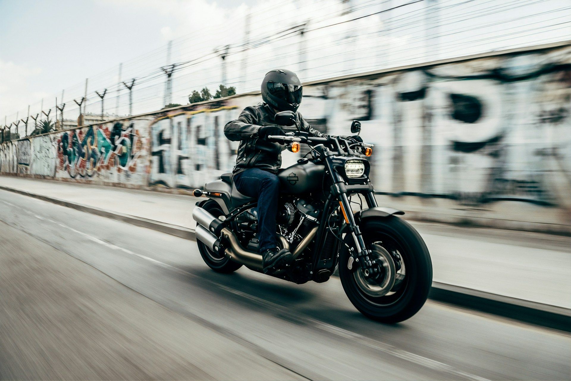 Black-clad motorcyclist riding a cruiser on a city street beside a graffiti-covered wall