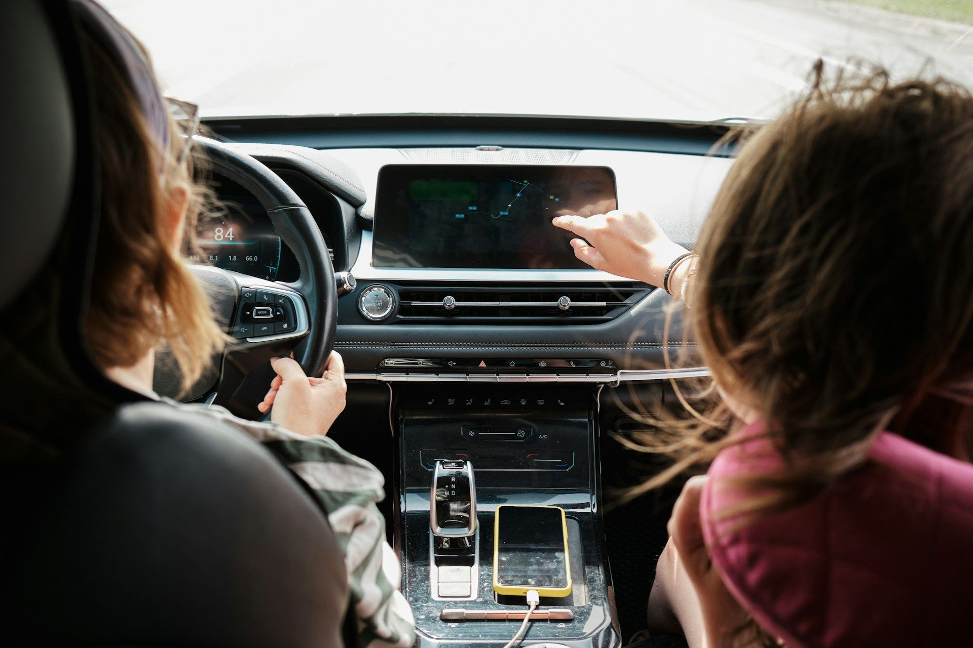 Two children in a car pointing at the dashboard touchscreen while driving on a road.