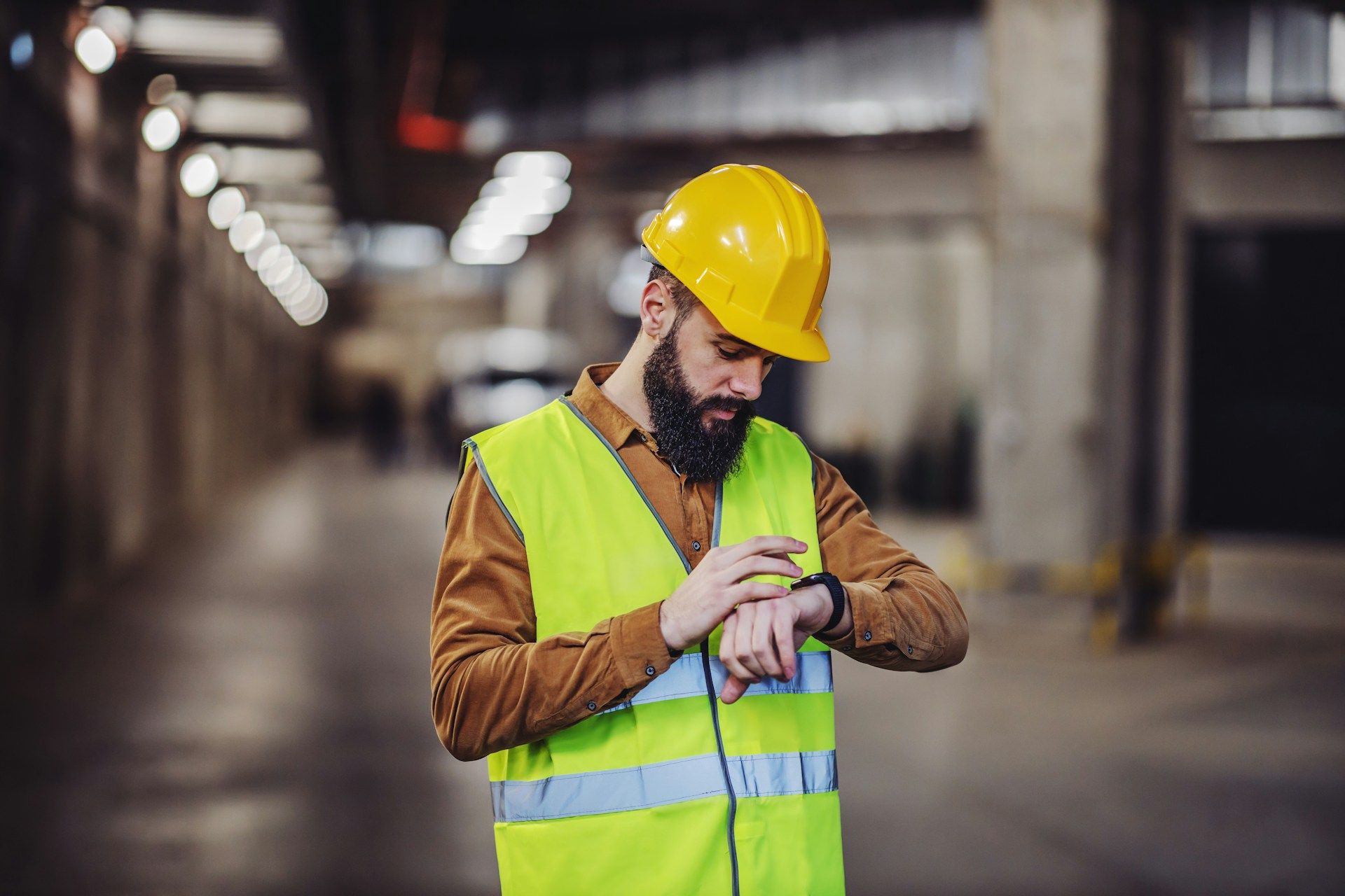 Worker in yellow hard hat and safety vest checking a wristwatch in an industrial warehouse