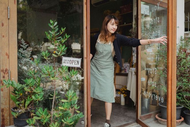 Woman opening a glass shop door, standing among potted plants and handwritten signs.