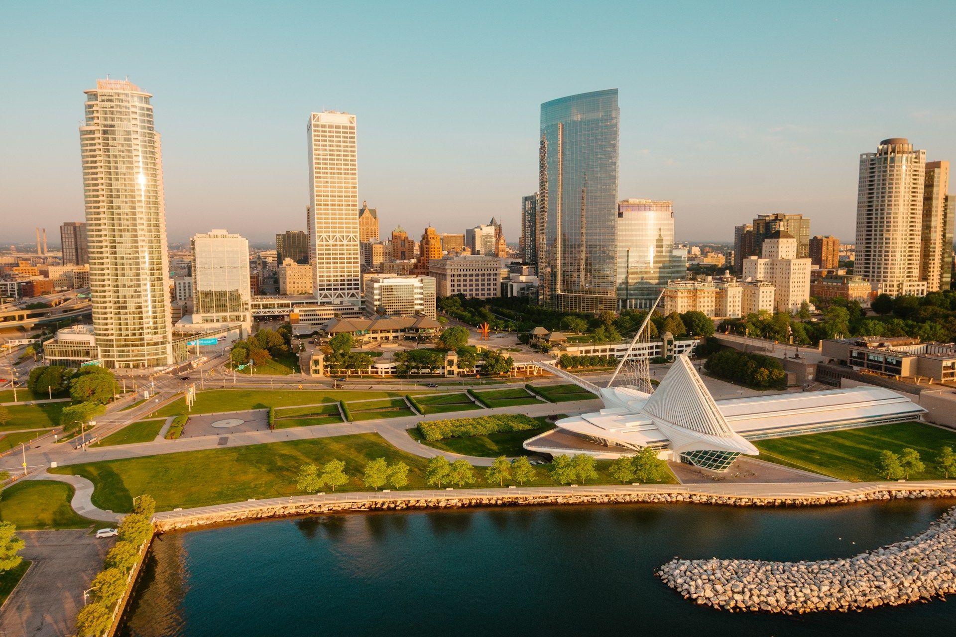 Modern waterfront park with white sculpture, green lawns, and city skyline at sunset
