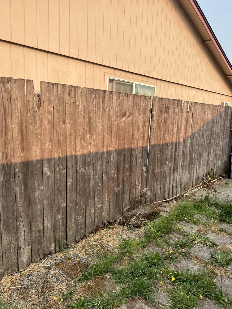 Weathered wooden fence beside a tan house, with patchy grass and dirt in the foreground.