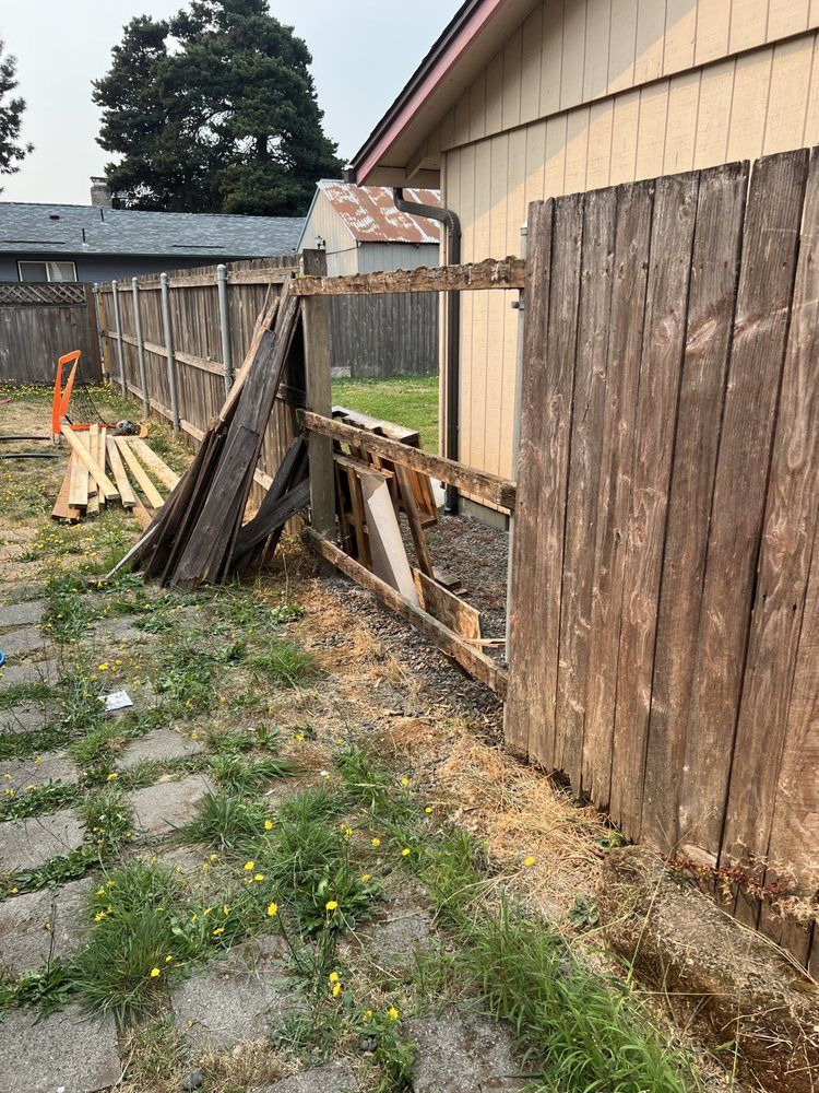 Collapsed wooden fence beside a house, with broken panels leaning into a backyard path.