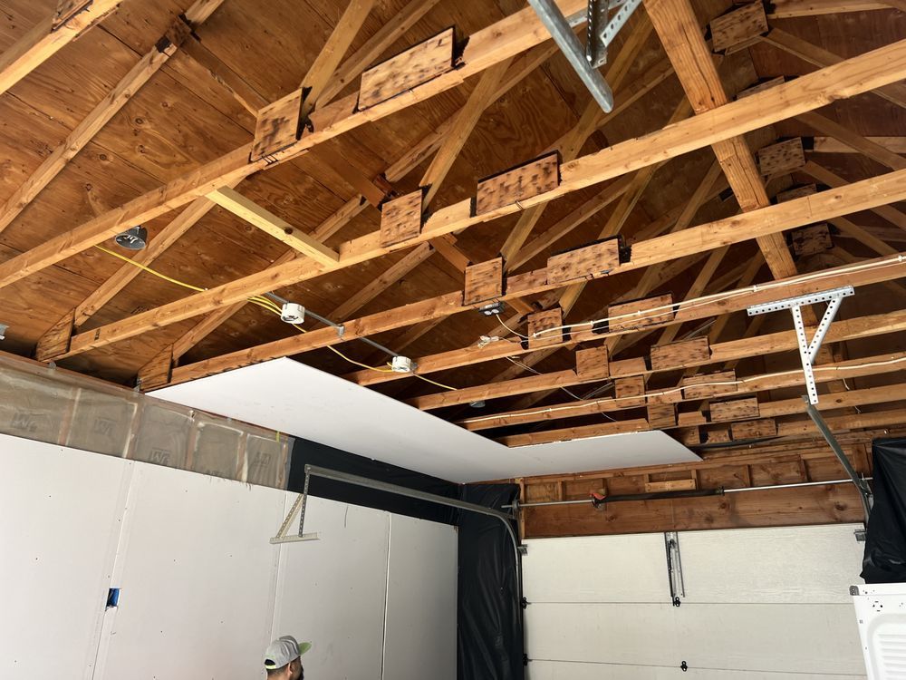 Unfinished garage ceiling with exposed wooden trusses, hanging lights, and white walls below.