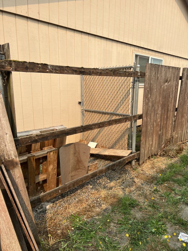 Weathered wooden fence panel leaning beside a chain-link gate against a tan building exterior.
