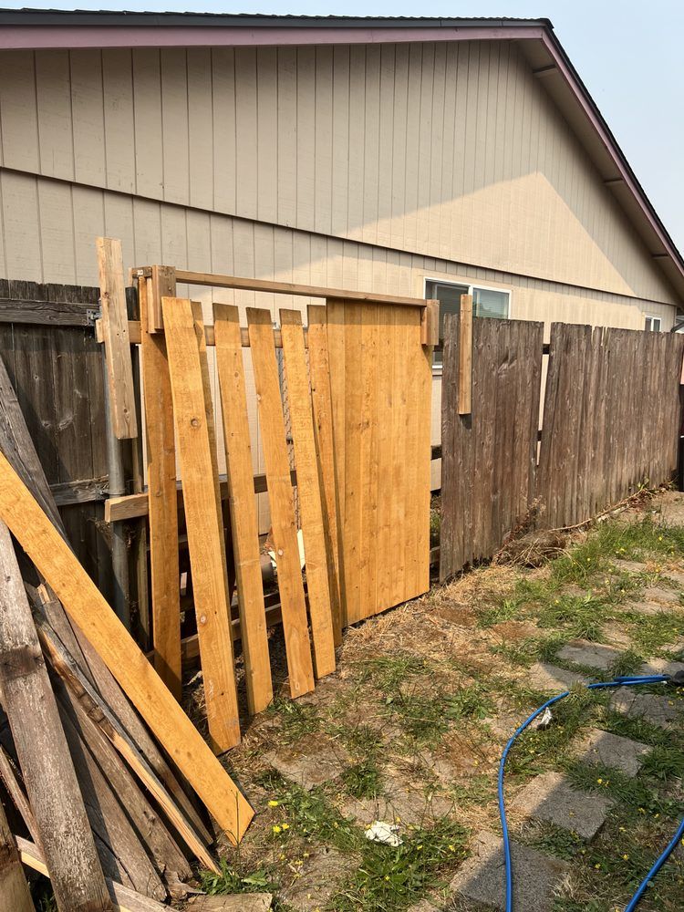Wooden fence panels leaning against a weathered fence beside a beige house and grassy yard.
