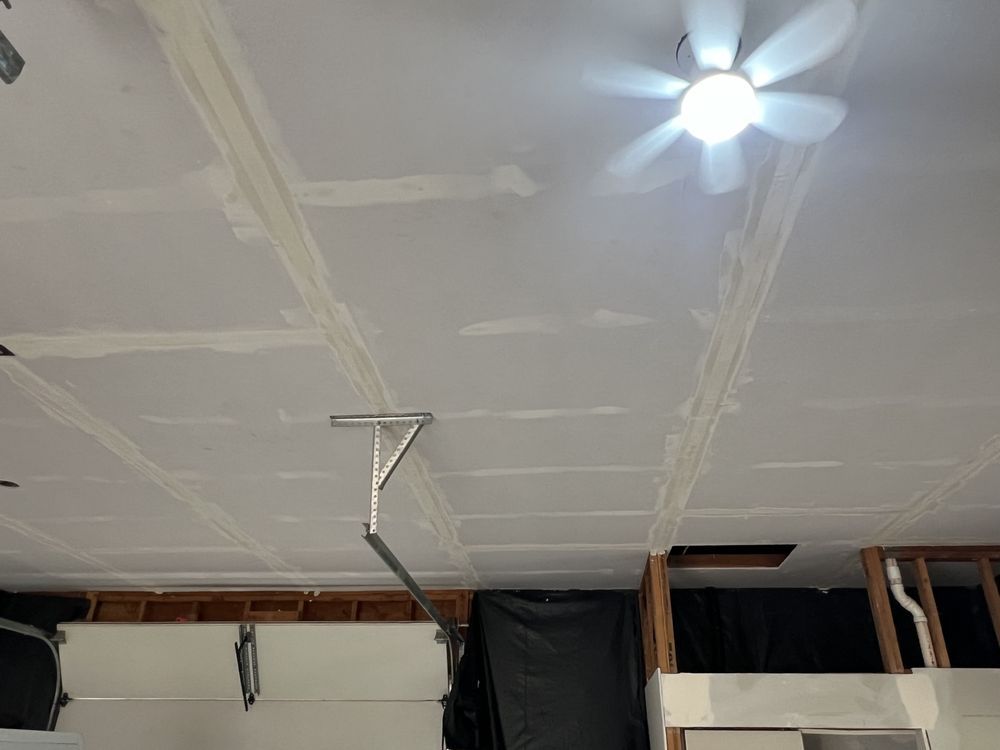 Partially finished ceiling with white drywall seams, a hanging wire, and a lit ceiling fan on the right