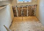 Bathroom gutted to the studs, with exposed wall framing and debris on the floor.