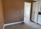 Empty room with tan walls, light wood floor, and a doorway beside white kitchen cabinets.