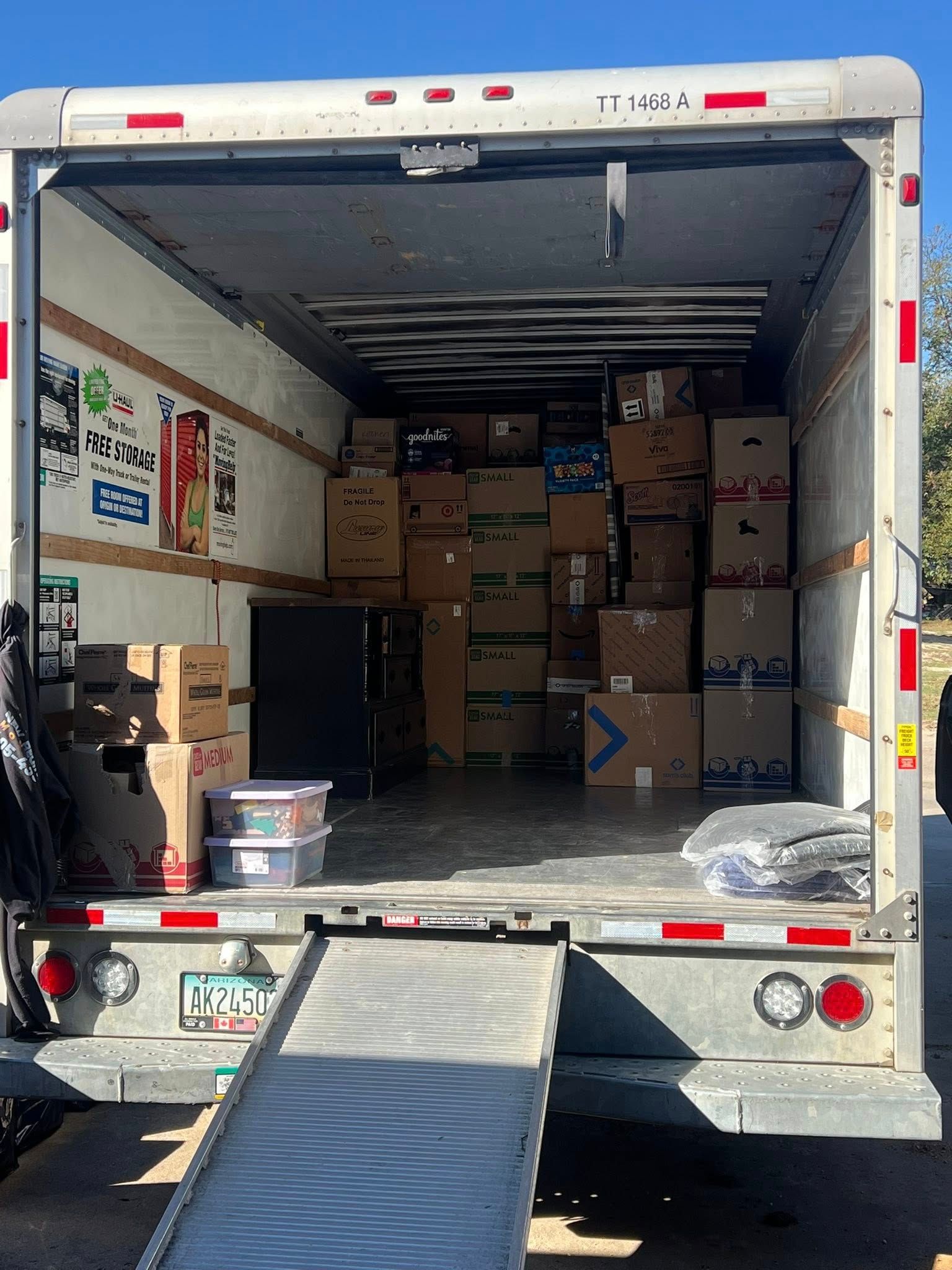 Open box truck loaded with stacked cardboard boxes, rear ramp down at a loading dock.