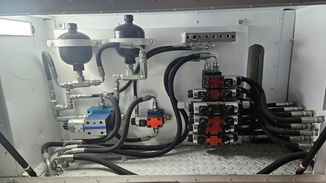 Maquinaria industrial con mangueras negras, válvulas rojas y tanques metálicos dentro de un armario de equipos blanco.