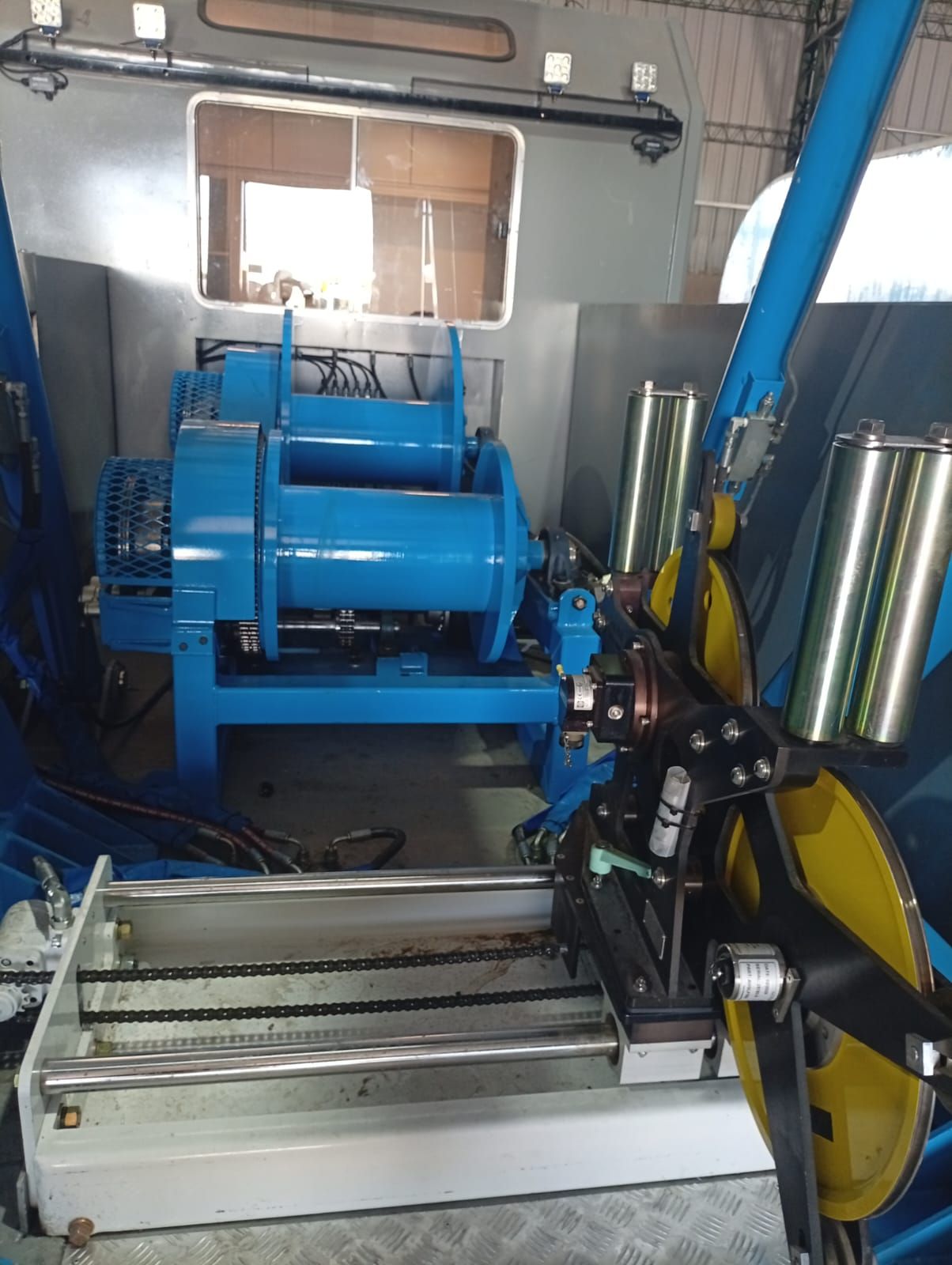Máquina industrial azul en un taller, con rodillos, controles y ruedas amarillas cerca de una ventana.