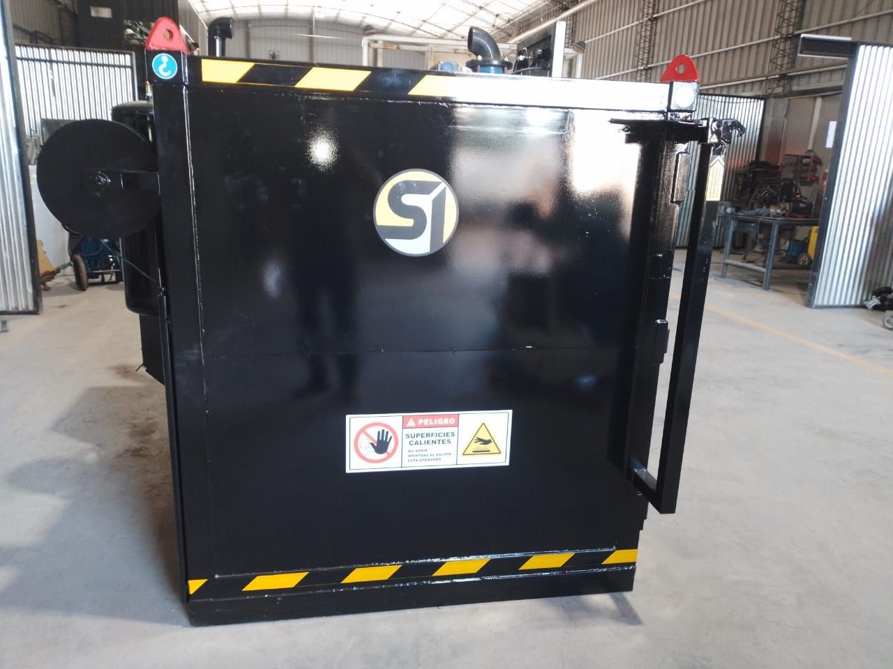 Máquina industrial negra con etiquetas de advertencia en un taller/almacén.