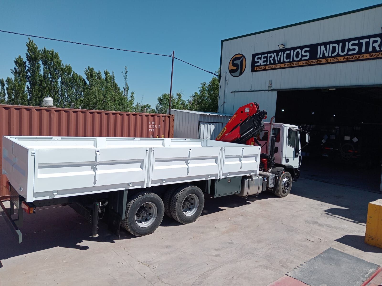 Camión volquete blanco con una grúa roja estacionado afuera de un garaje industrial bajo un cielo azul despejado.