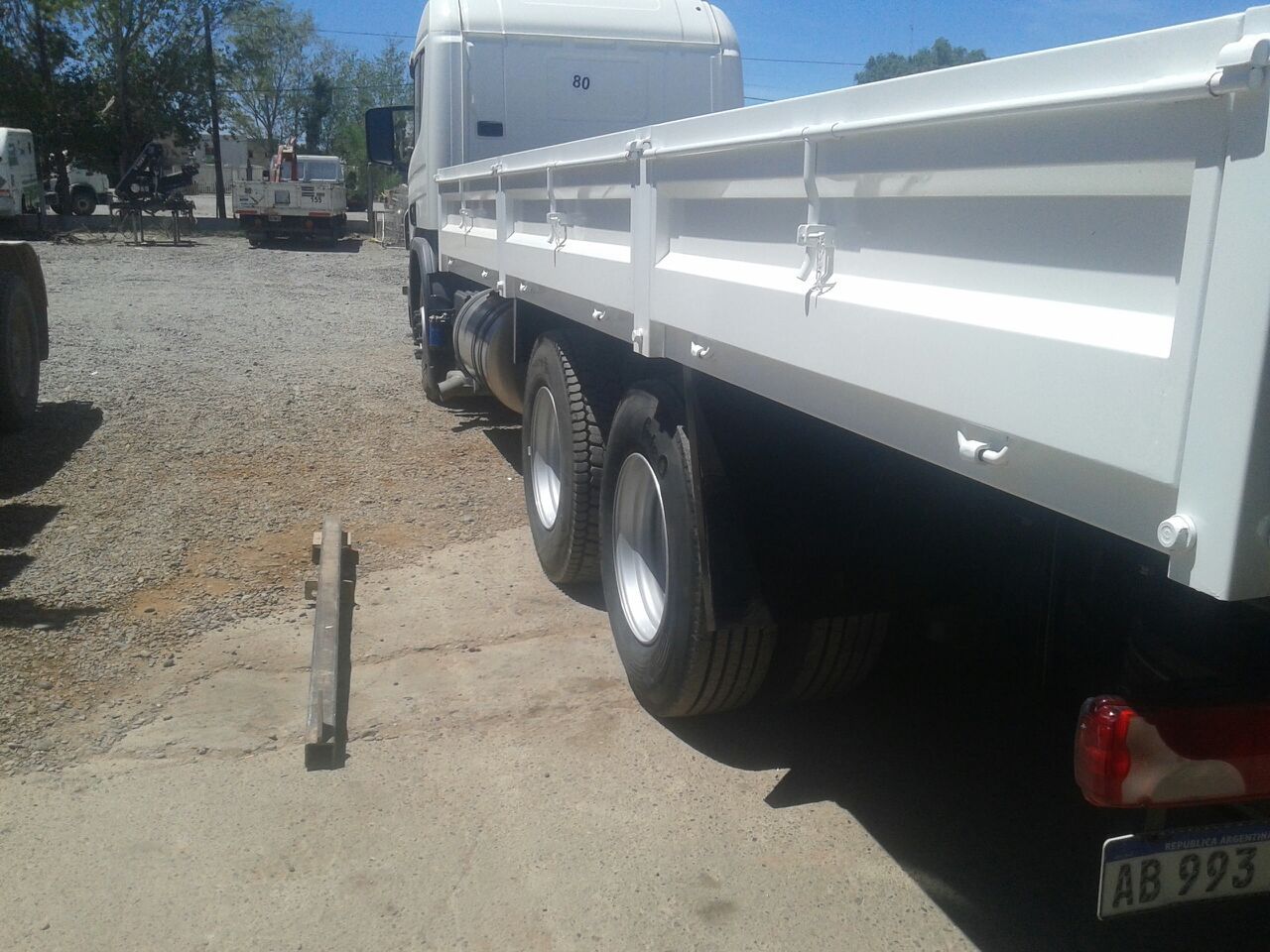 Camión blanco de plataforma plana estacionado sobre grava junto a un camino de entrada bajo la luz del sol.