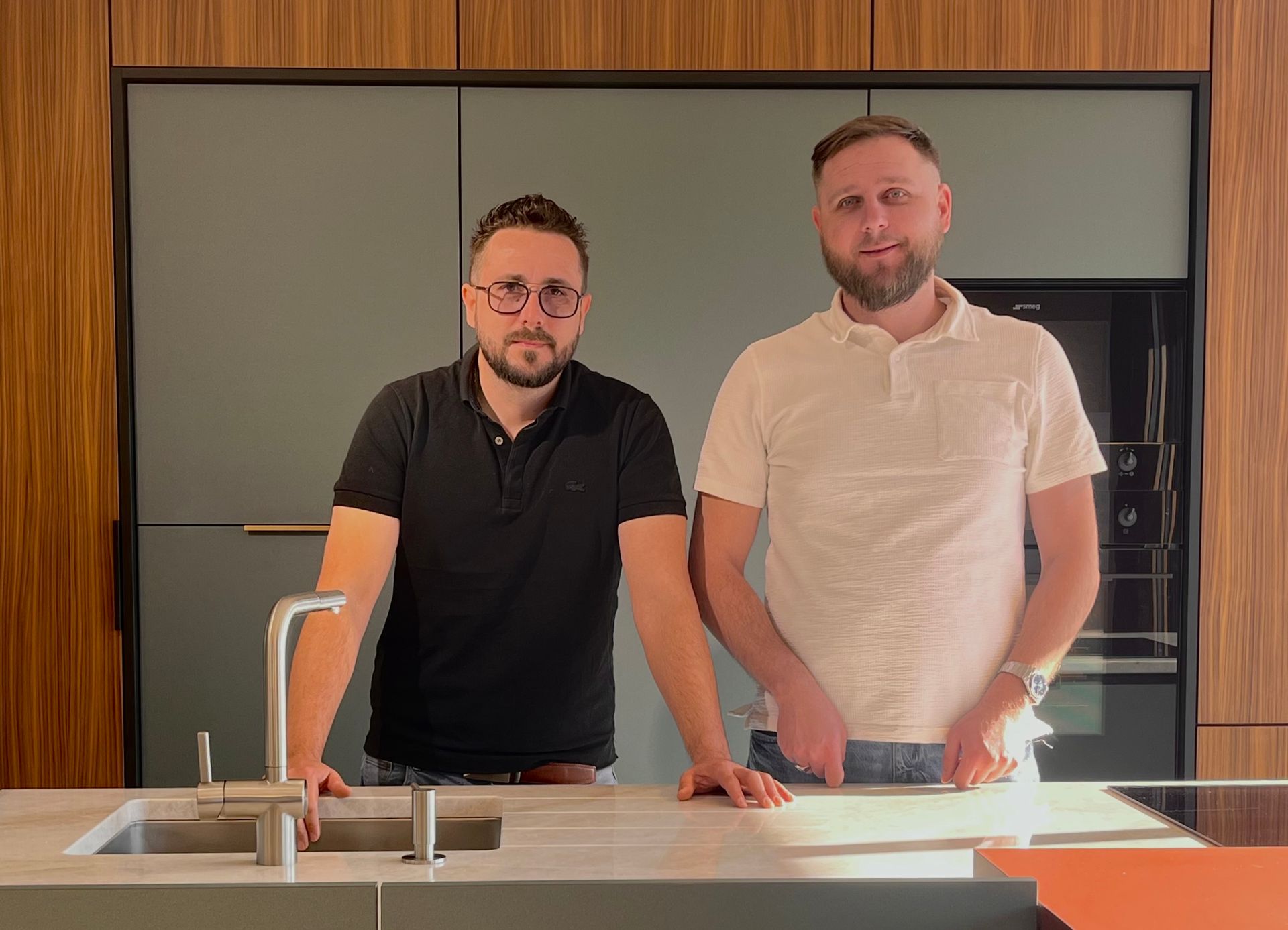 Deux hommes se tiennent derrière un îlot de cuisine dans une cuisine moderne, l'un en chemise noire et l'autre en chemise blanche.