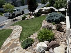 Landscaped front yard with curved stone walkway, green lawn, rocks, and shrubs beside a residential street