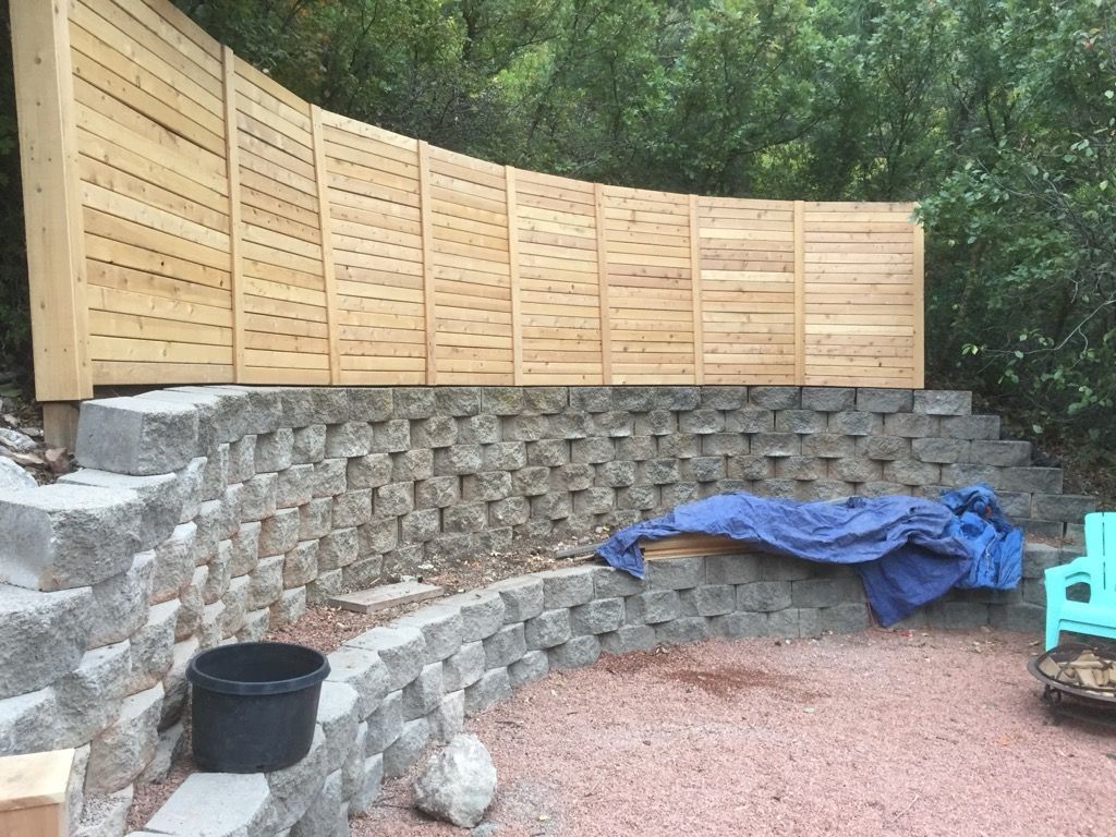 Wooden privacy fence atop a stone retaining wall, with a gravel yard and blue tarp in a wooded backyard.