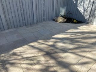 Empty paved patio beside a corrugated metal fence, with a bag in the corner and tree shadows across the ground