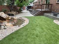 Backyard patio with green lawn, stone garden bed, and wooden deck steps beside a brick house