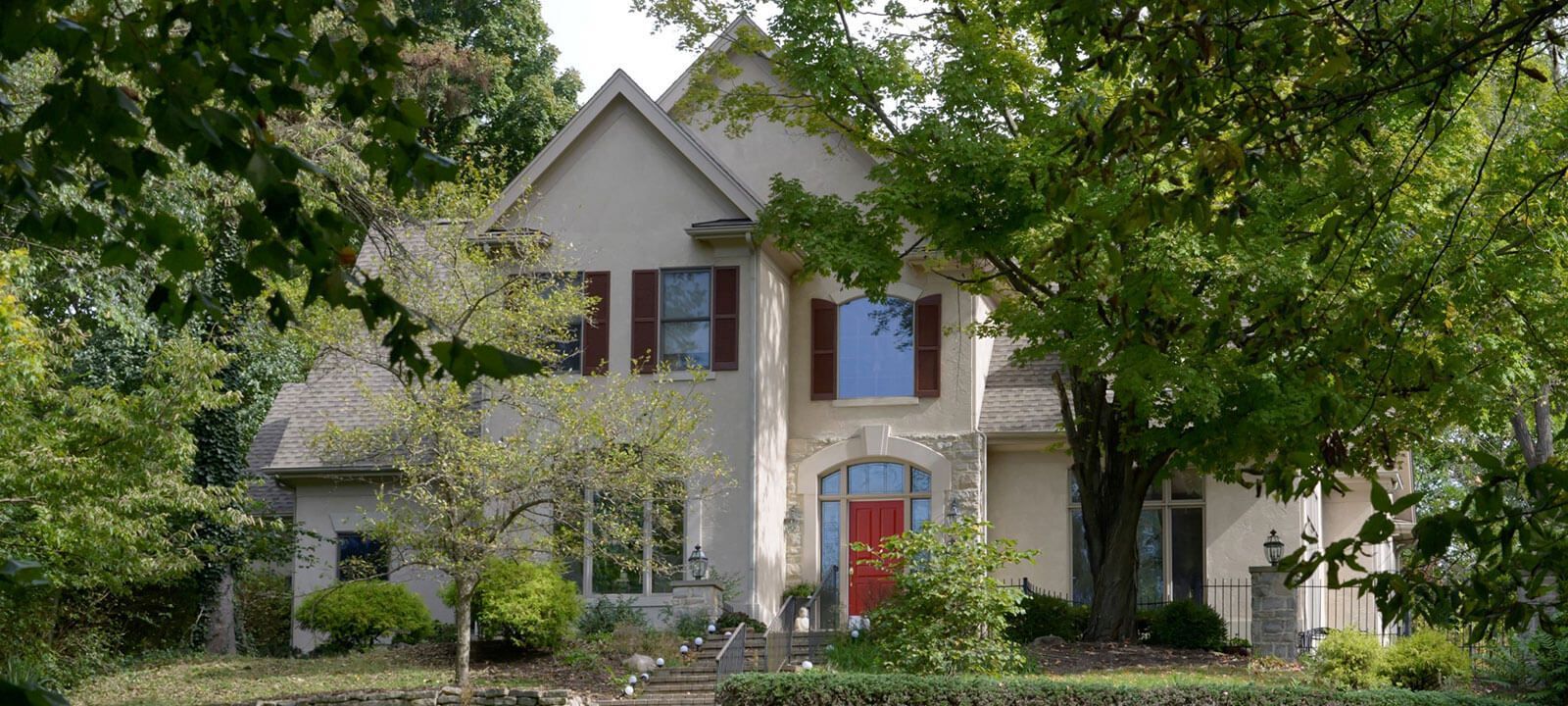 Two-story house with red door, stone archway, and brown shutters, framed by green trees and foliage.