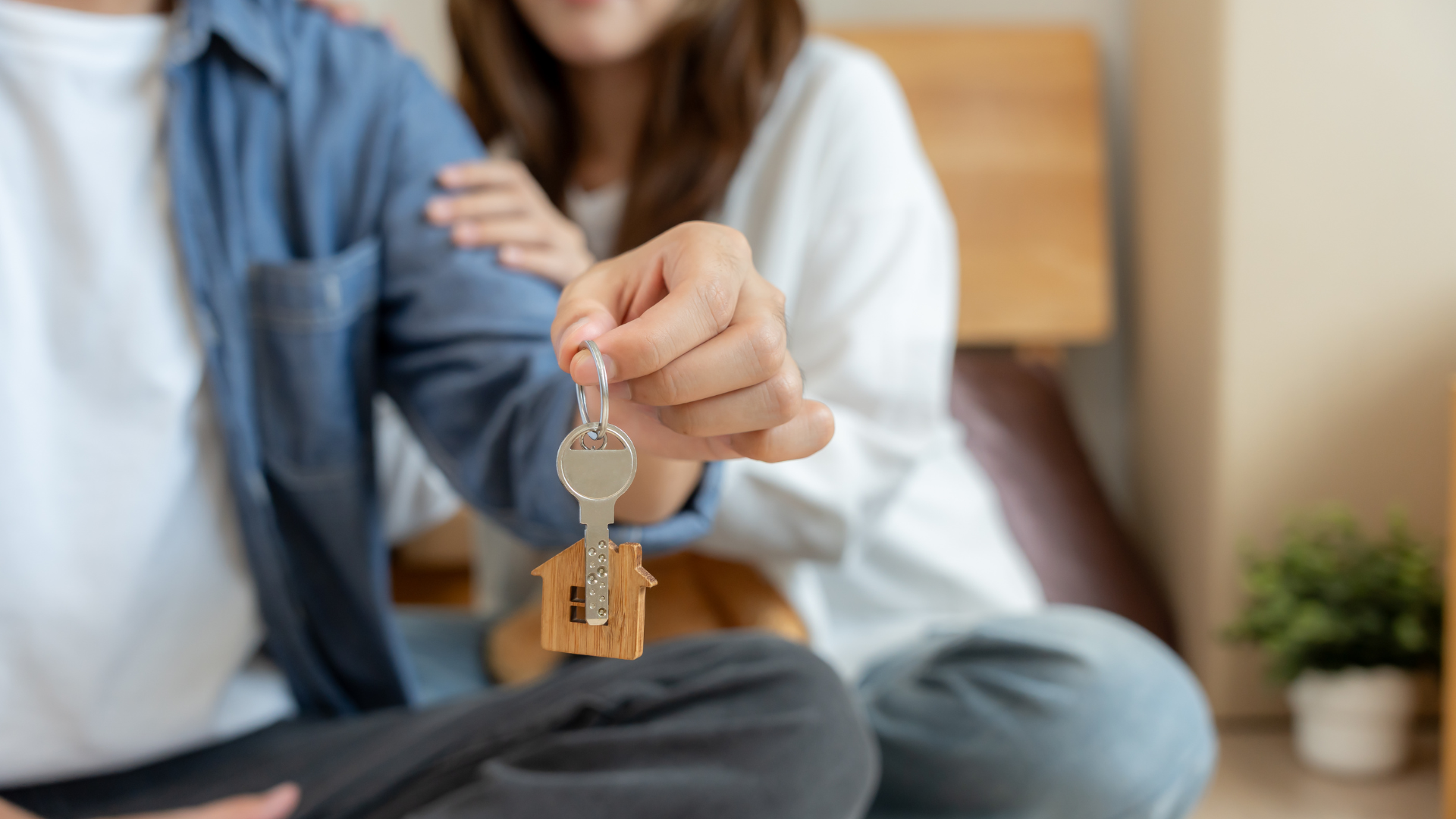 Person holding house key with wooden house charm. Another person in background.