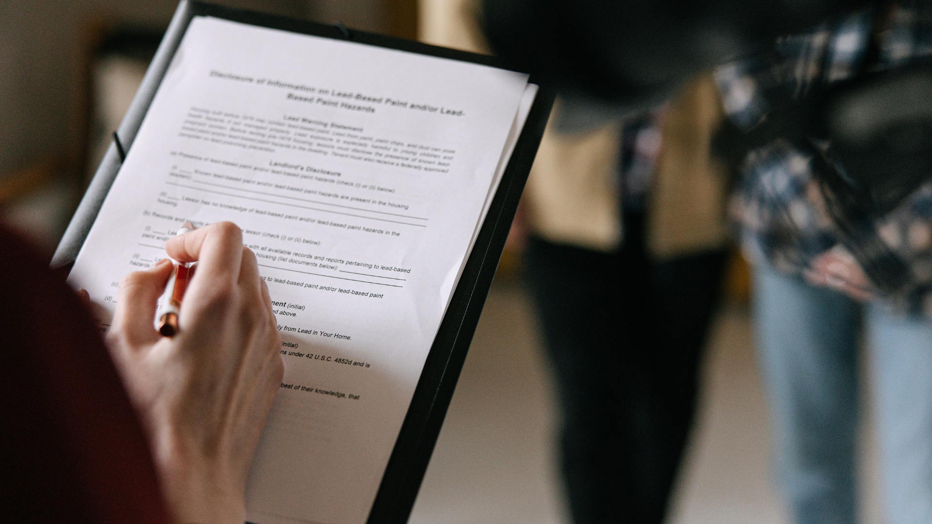 Person writing on document held in a clipboard, with two other people in background.