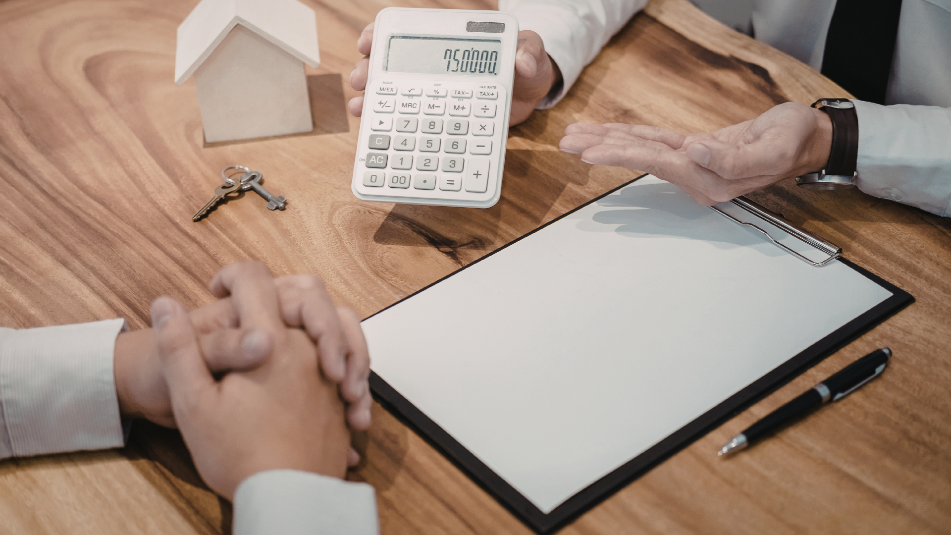 Hands clasped at a table during a meeting with a calculator, house model, keys, and paperwork.