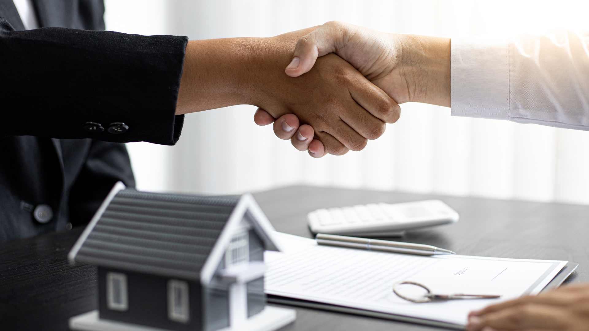 Two people shaking hands over a miniature house, documents, and keys, likely sealing a real estate deal.