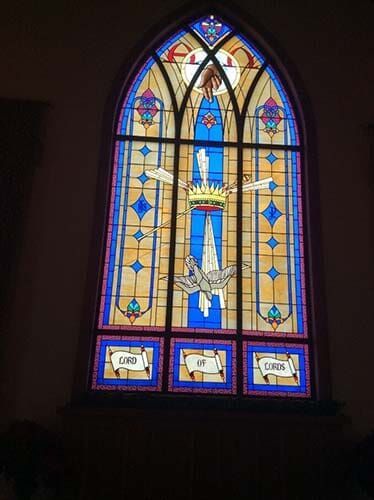 Stained glass church window with blue, yellow, and white colors, depicting a dove, crown, and clouds.