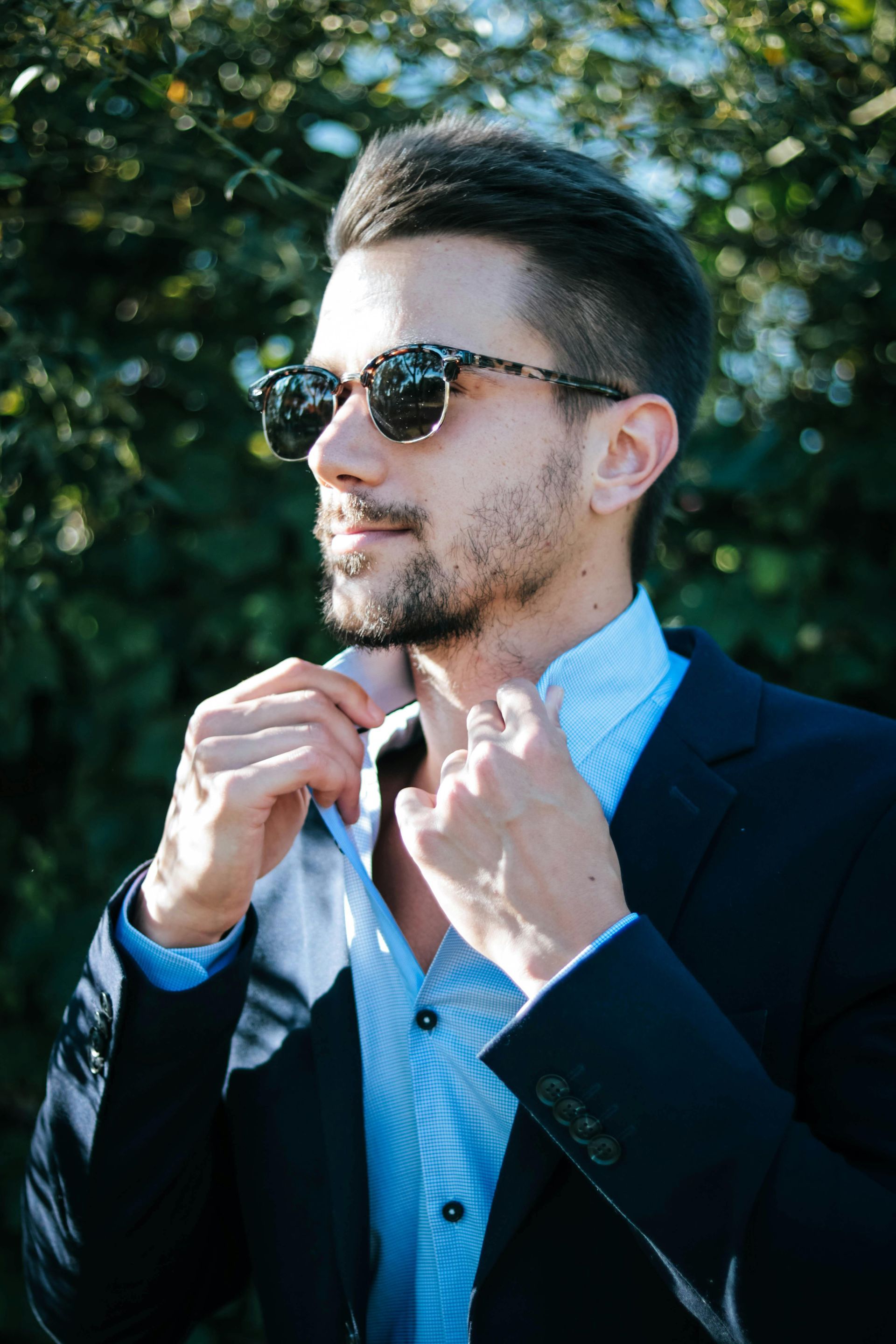Man in sunglasses adjusts his shirt collar, wearing a blue suit jacket, in front of green foliage.
