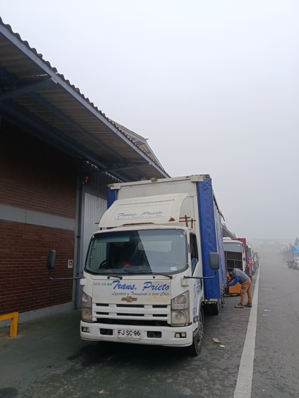 Camión blanco estacionado junto a una zona de carga de un almacén en un día de niebla, con otro camión detrás.