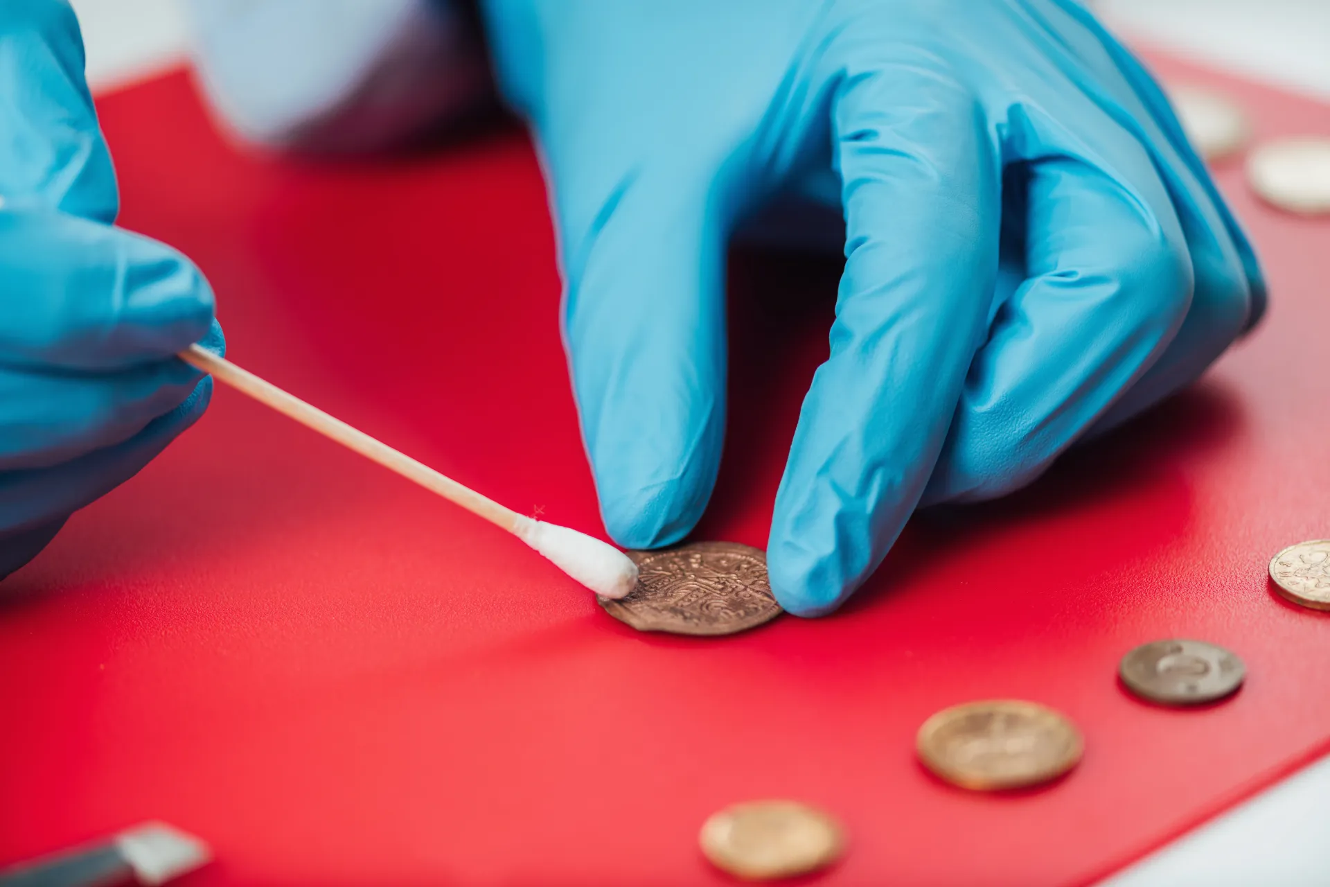 Gloved hands cleaning a coin with a cotton swab on a red surface
