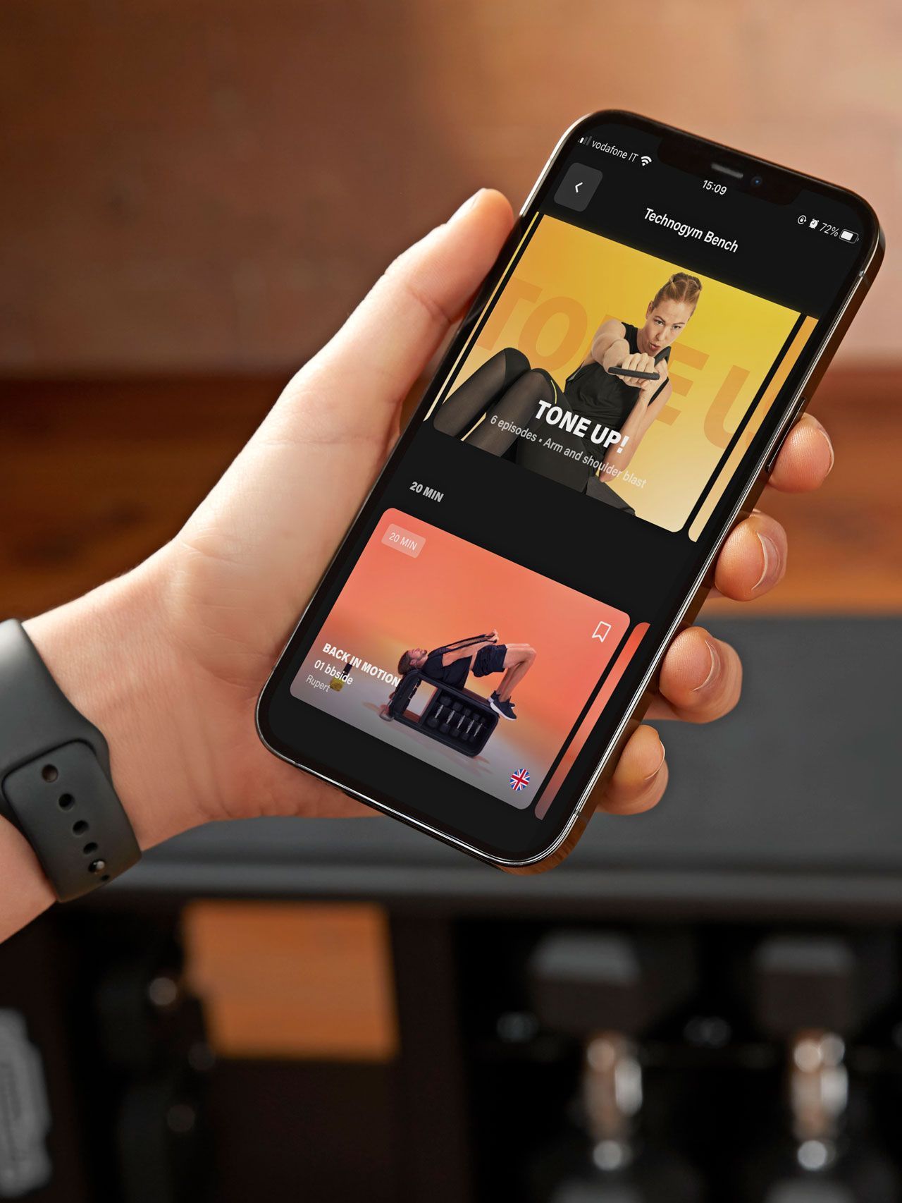 A hand holds a smartphone displaying a fitness app interface with workout video thumbnails against a blurred gym background.