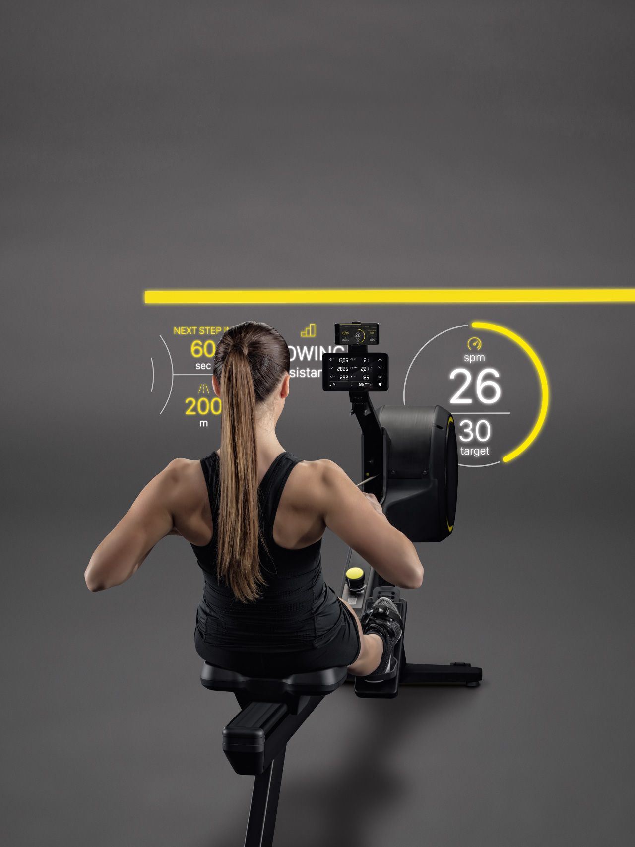 A person uses a rowing machine in a studio, facing a digital display showing workout metrics like pace and stroke rate.