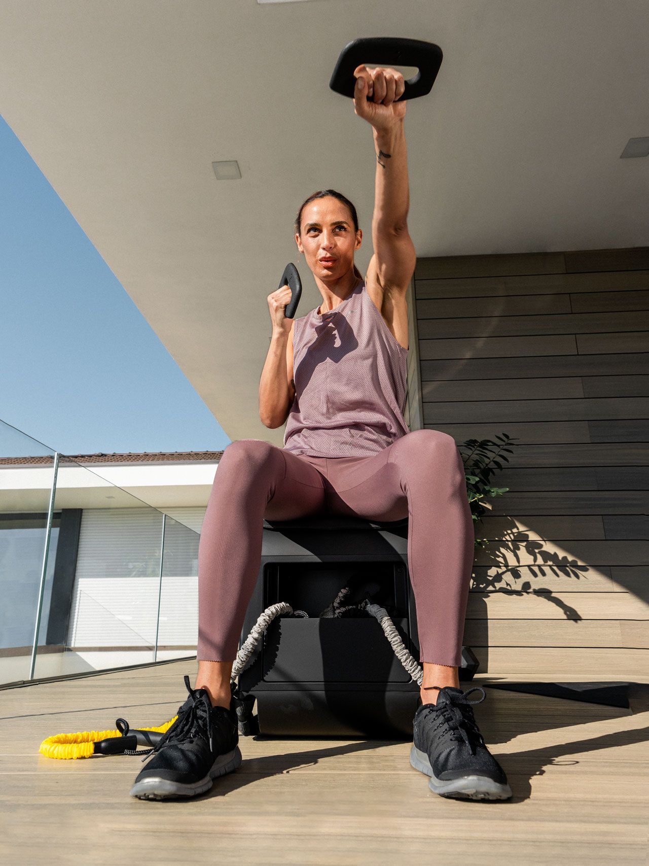 A person sits on a fitness box outdoors, performing a one-arm overhead press with a kettlebell, wearing mauve activewear.