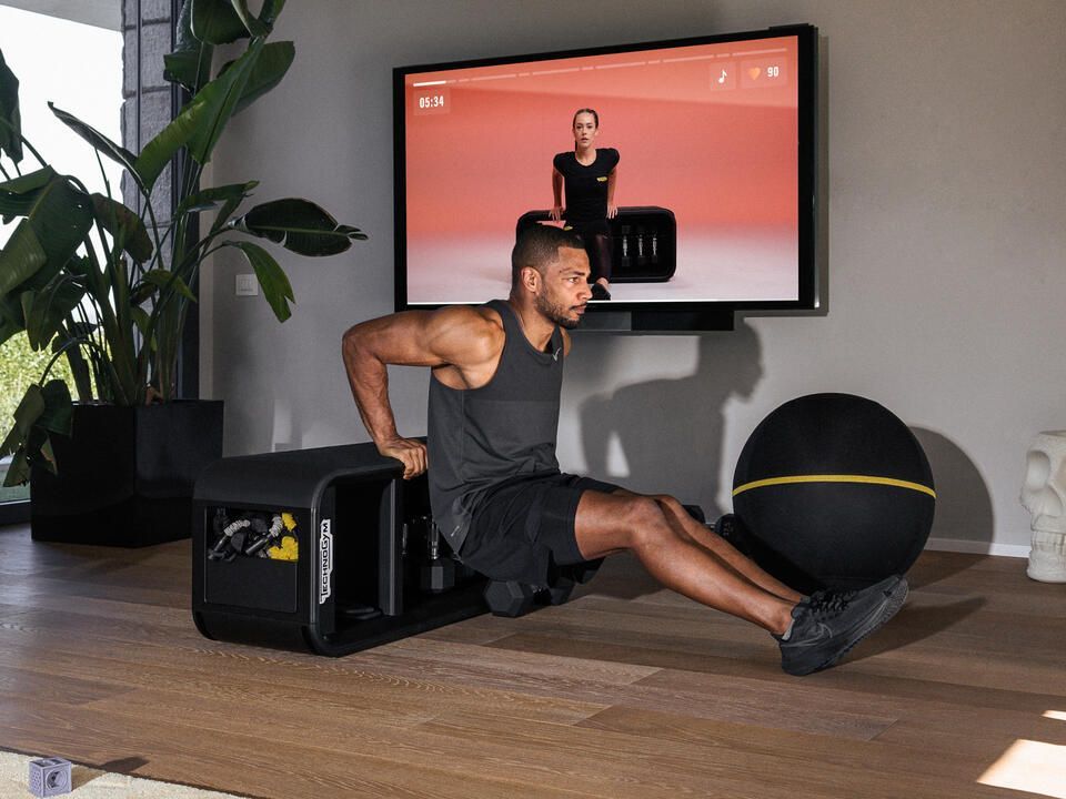 A person performs tricep dips on a home gym bench in a modern room with a large screen displaying a workout guide.