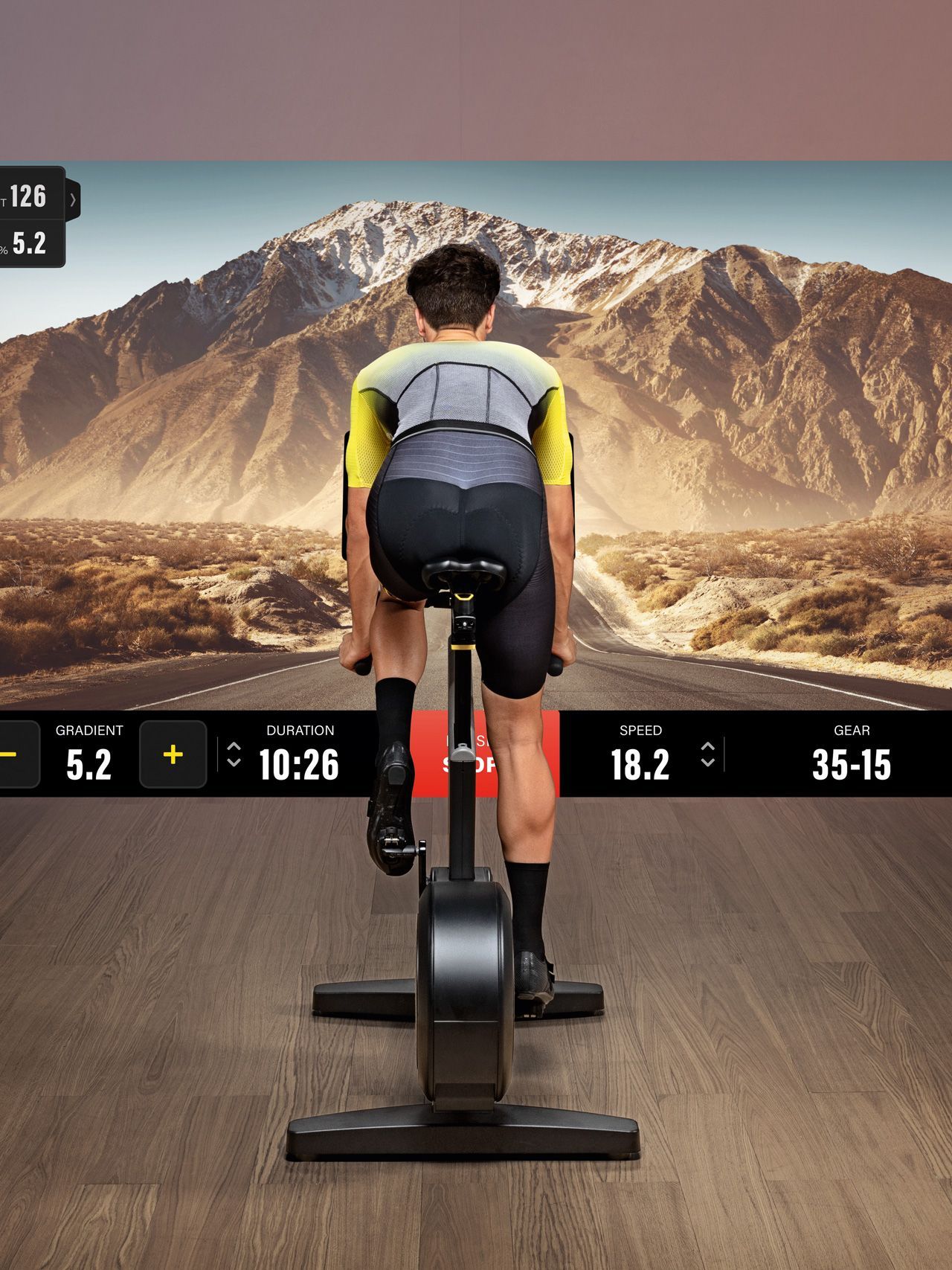 A cyclist rides a stationary bike in front of a screen displaying a mountain road, with digital performance metrics overlaid.