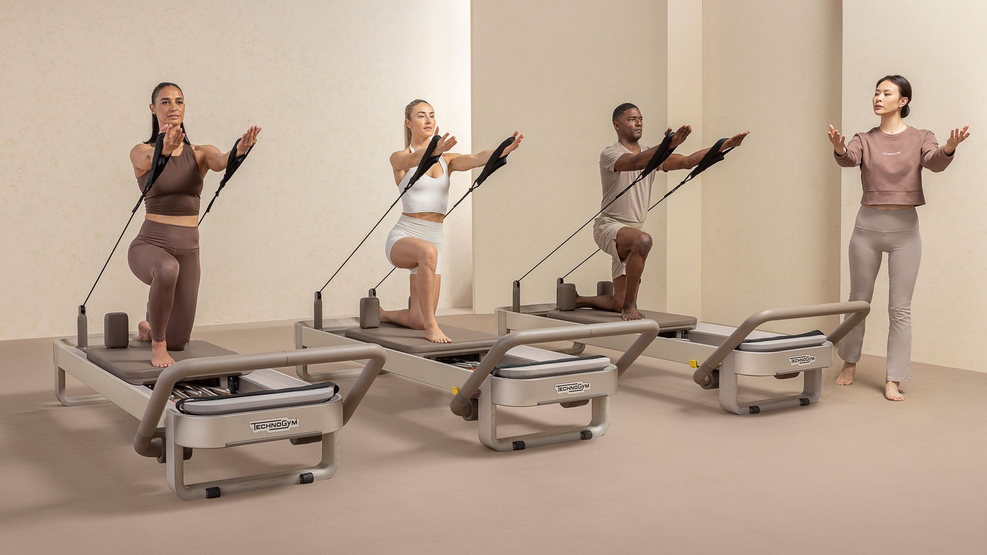 Three people kneel on Pilates reformers using resistance straps while an instructor leads them in a bright studio.