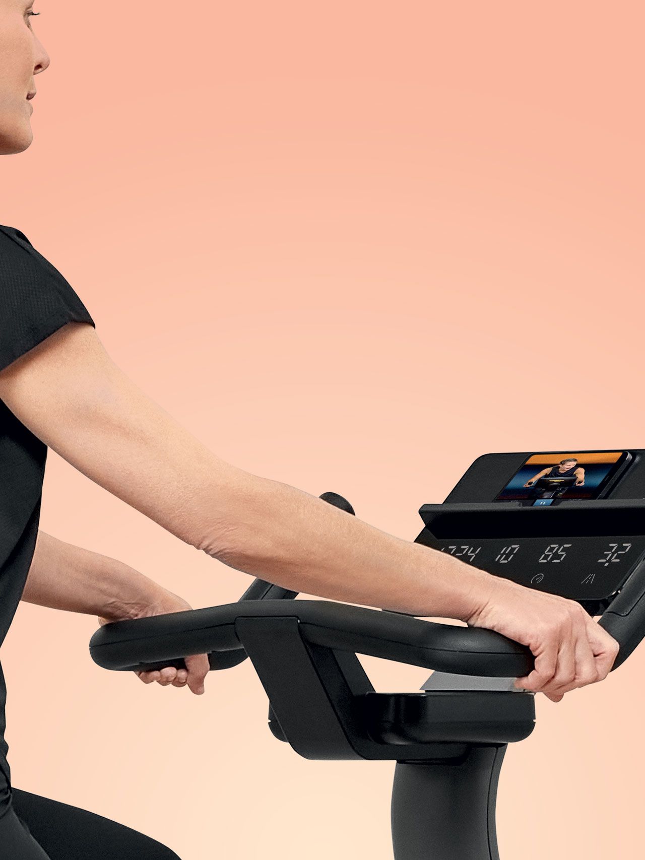 A person in black workout clothing holding the handlebars of an indoor exercise bike against a solid peach background.