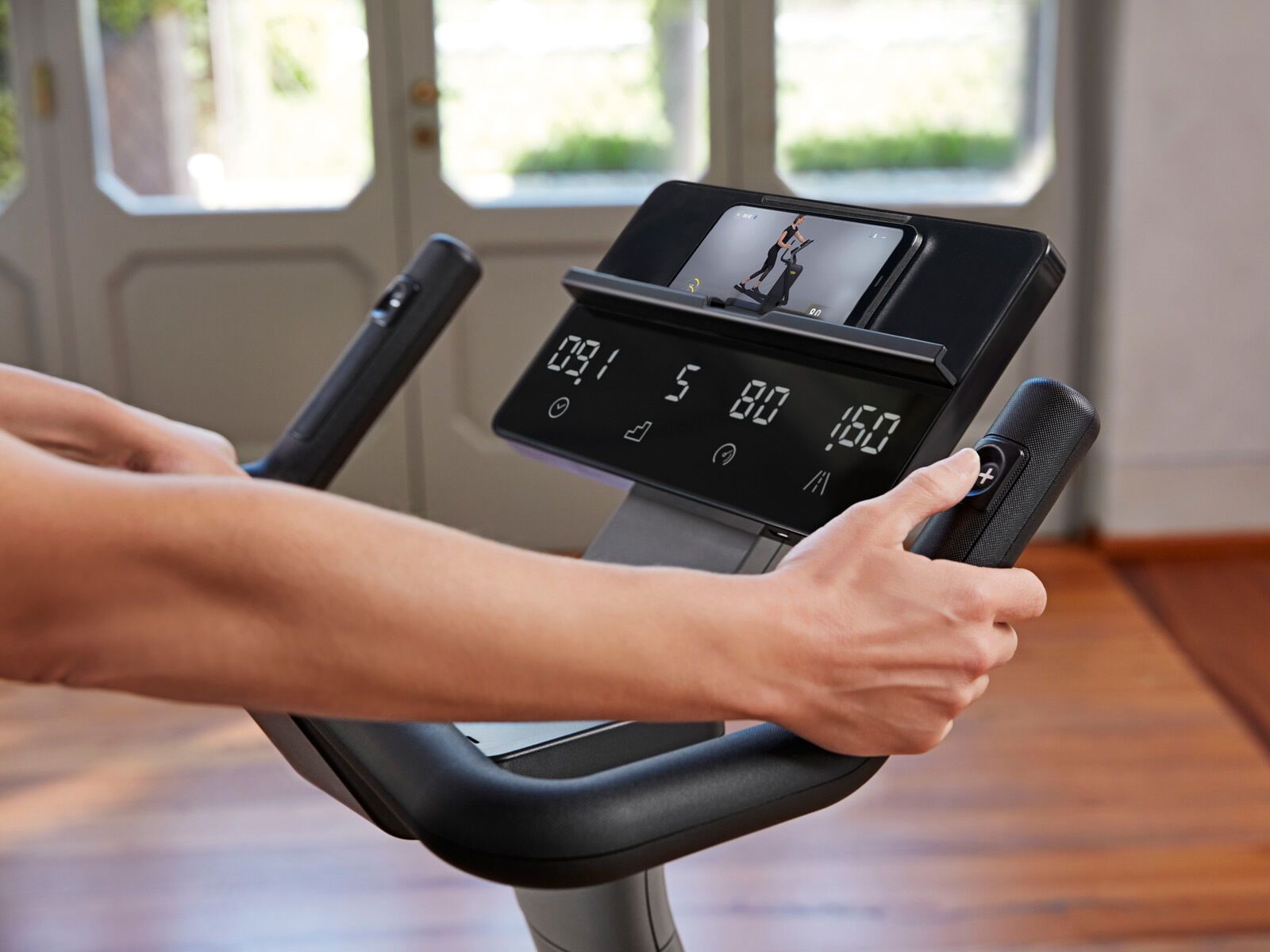 Close-up of hands on a stationary bike handlebar console displaying workout data with a phone held in the mount.