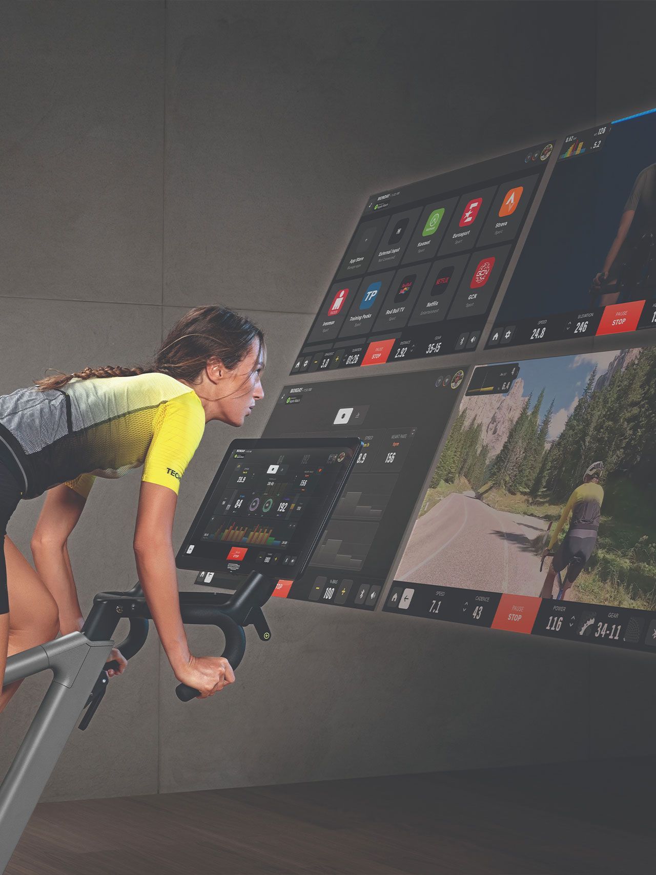 A person cycles on an indoor stationary bike facing a large, multi-paneled display showing workout data and scenery.