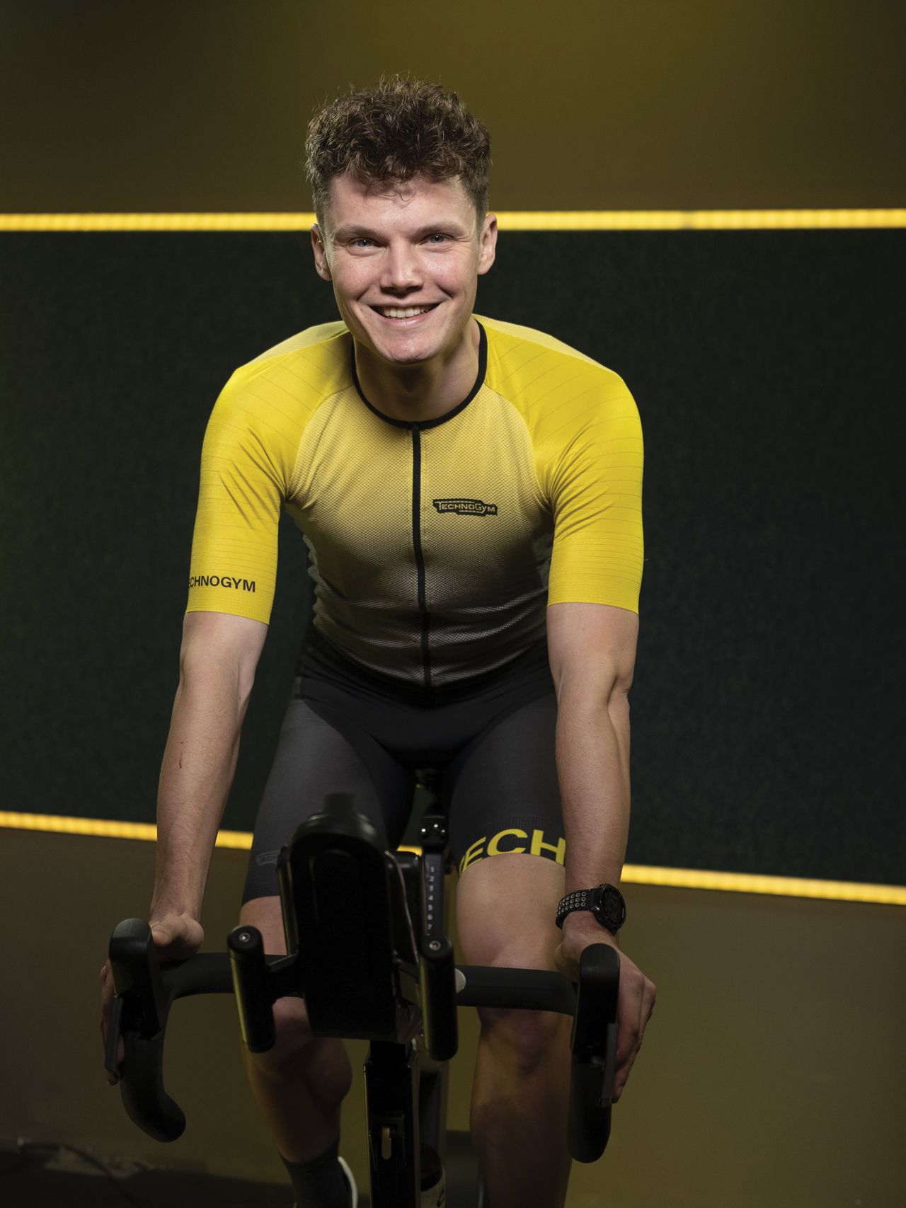 A person in a yellow and black cycling jersey smiles while sitting on a stationary exercise bike in a dark studio.