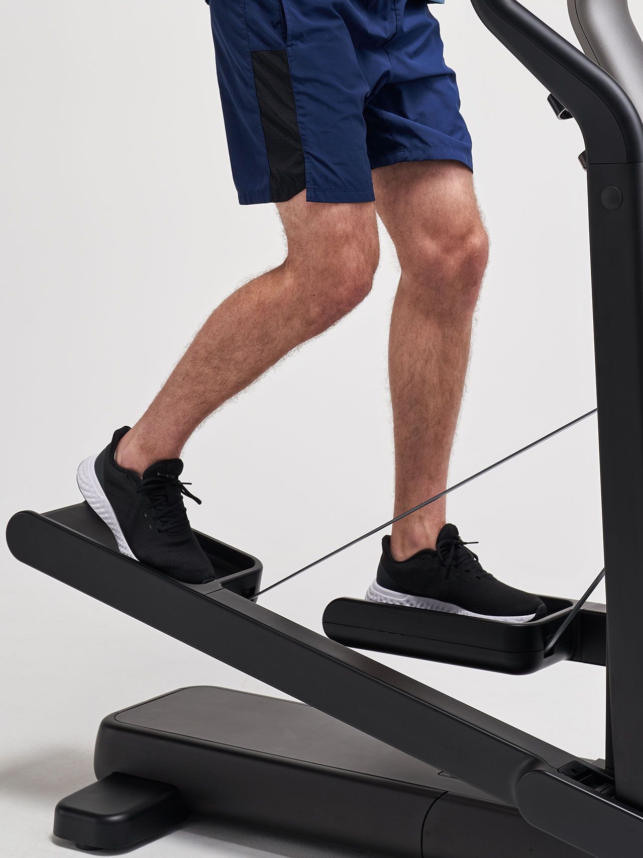 Lower body of a person in navy shorts and black sneakers exercising on a black stepper machine.