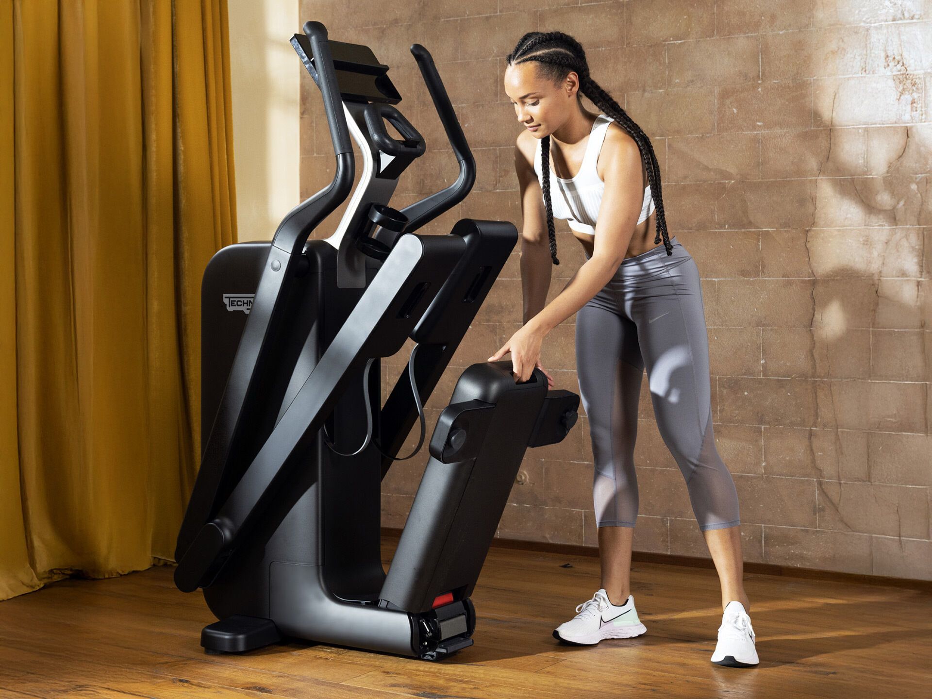 A person folds a black Technogym elliptical machine in a room with a wood floor and textured wall.