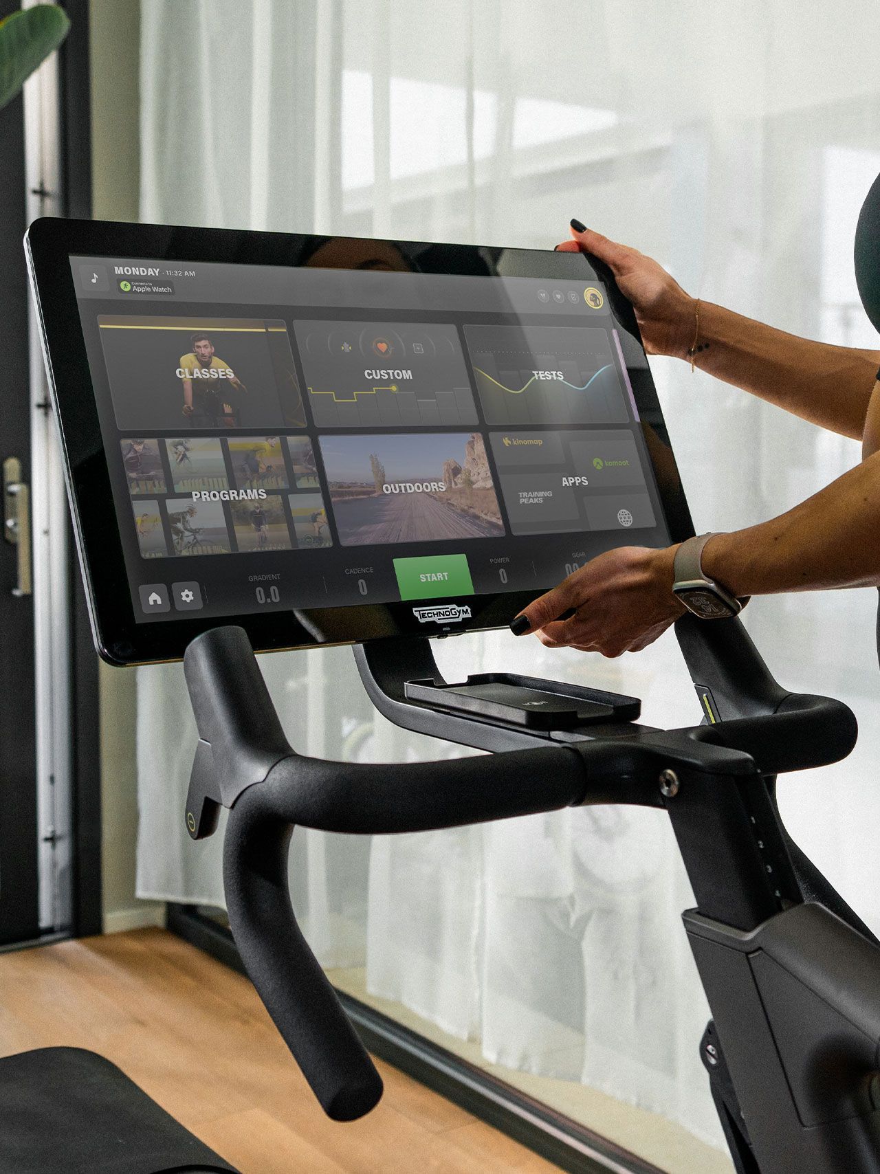 A person interacting with the touchscreen display of a Technogym stationary bike in a bright, modern room.