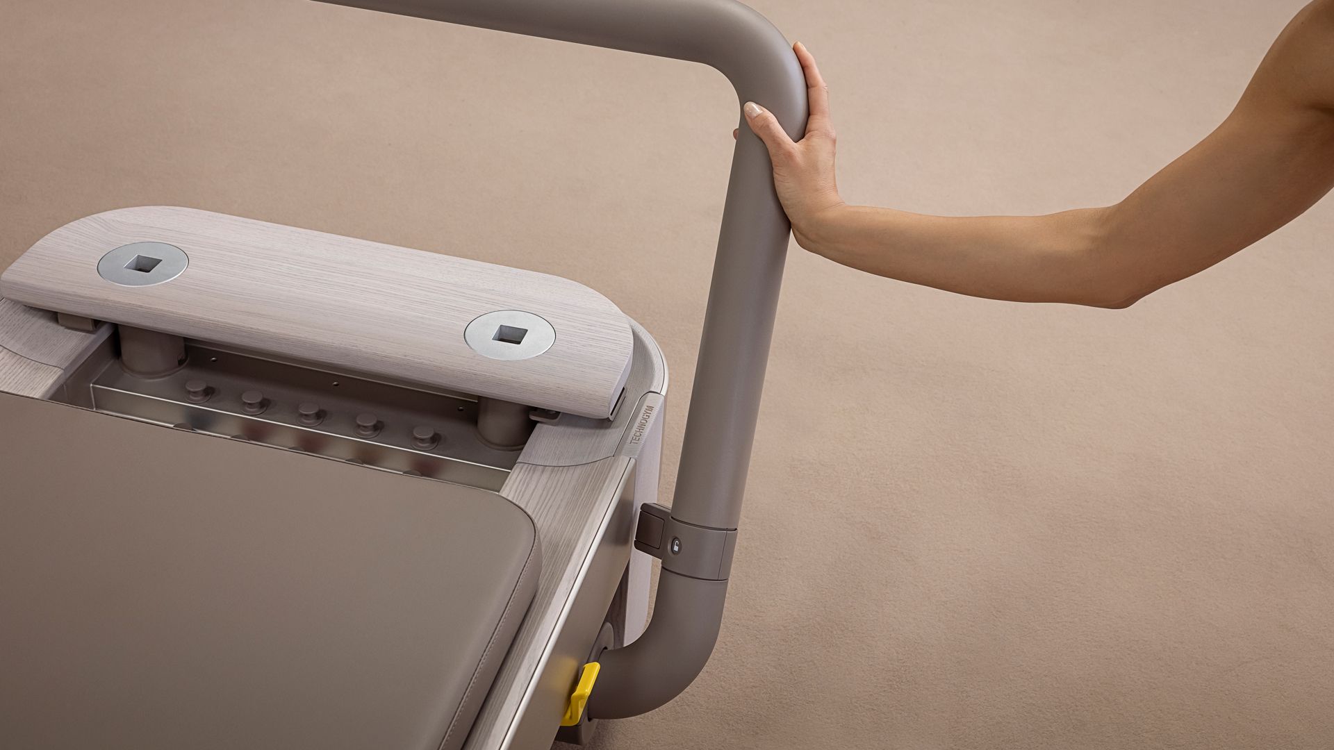 A person holds the gray handle of a piece of exercise equipment, likely a Pilates reformer, on a neutral-colored floor.