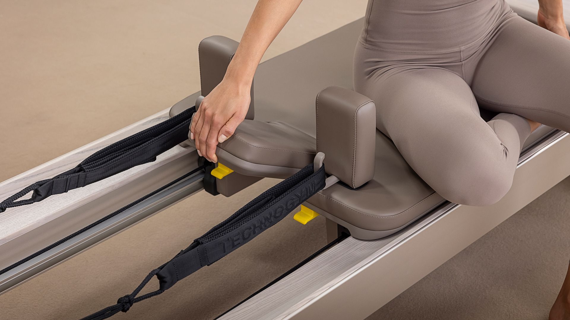 A person sits on a neutral-toned Pilates reformer machine, their hand resting on the adjustable shoulder rest.