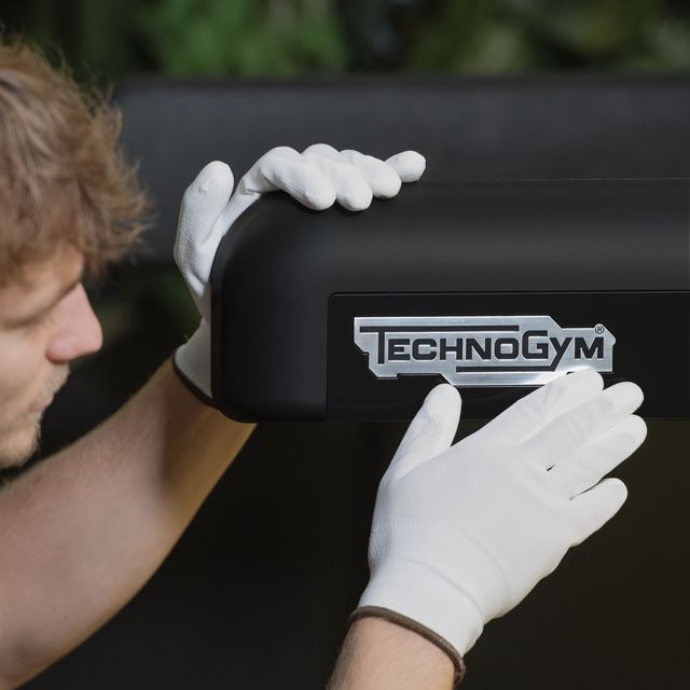 A person wearing white cotton gloves carefully touches a silver Technogym logo on black gym equipment.